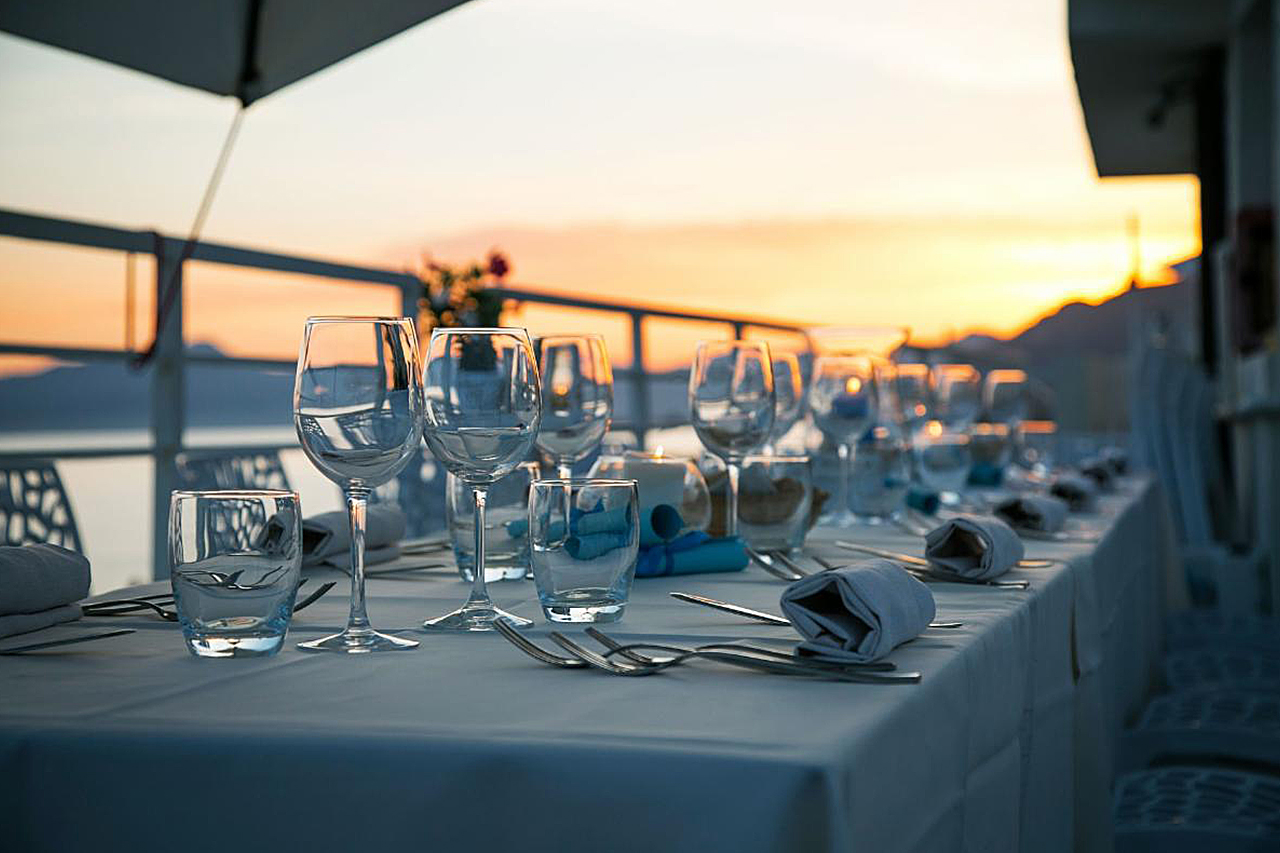 Elegant outdoor dinner table set with glassware and white linens at sunset, overlooking a scenic waterfront view.